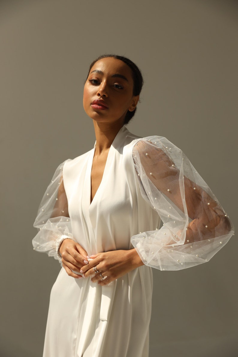 May include: A white satin robe with sheer, pearl-embellished sleeves. The robe has a V-neckline and a tie belt.