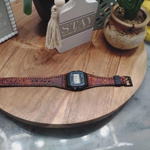 Può includere: Un orologio digitale nero con un cinturino in pelle marrone con cuciture decorative. L'orologio è esposto su un vassoio rotondo di legno. Il cinturino dell'orologio ha una fibbia nera.