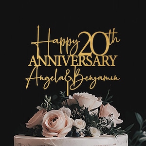 May include: A two-tiered white cake decorated with pink roses and greenery. A gold cake topper reads "Happy 20th Anniversary Angela & Benjamin" in elegant script. The cake sits against a black background.
