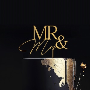May include: A two-tiered black wedding cake with gold leaf accents and dripping icing. A gold "MR & Mrs" cake topper is placed on top. The cake is set against a black background.