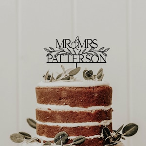 May include: A black and white wedding cake topper with the text "MR & MRS PATERSON" and a leafy design. The topper is on top of a multi-tiered cake with white frosting and brown cake layers.