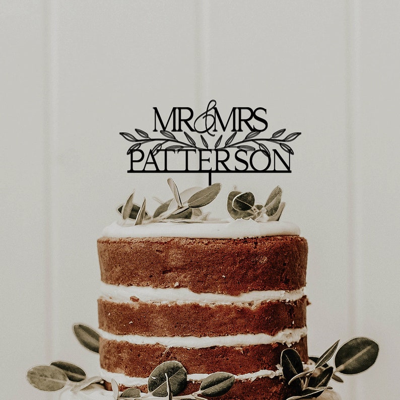 May include: A black and white wedding cake topper with the text "MR & MRS PATERSON" and a leafy design. The topper is on top of a multi-tiered cake with white frosting and brown cake layers.