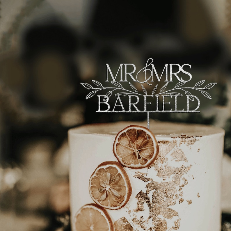 May include: A white wedding cake with gold leaf accents and dried orange slices. A silver cake topper reads "Mr. & Mrs. Barfield" with a leafy design.