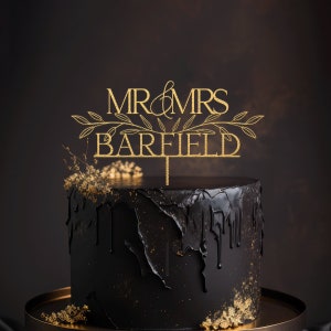 May include: Gold glitter wedding cake topper with the text "MR & MRS BARFIELD" and leafy branches on either side.
