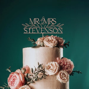 May include: A two-tiered wedding cake decorated with pink and white flowers. The cake topper says "Mr & Mrs Stevenson" in a script font.