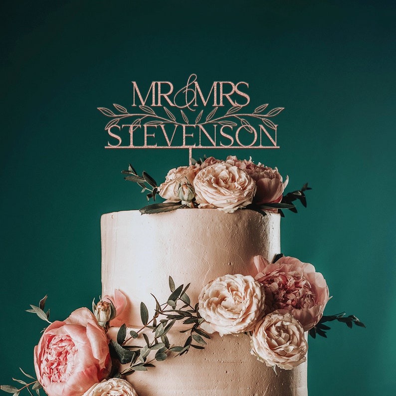 May include: A two-tiered wedding cake decorated with pink and white flowers. The cake topper says "Mr & Mrs Stevenson" in a script font.