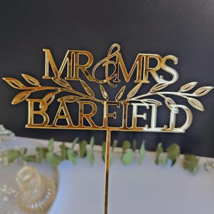 May include: Gold mirror acrylic cake topper with the text "MR & MRS BAREFIELD" and a leafy design.