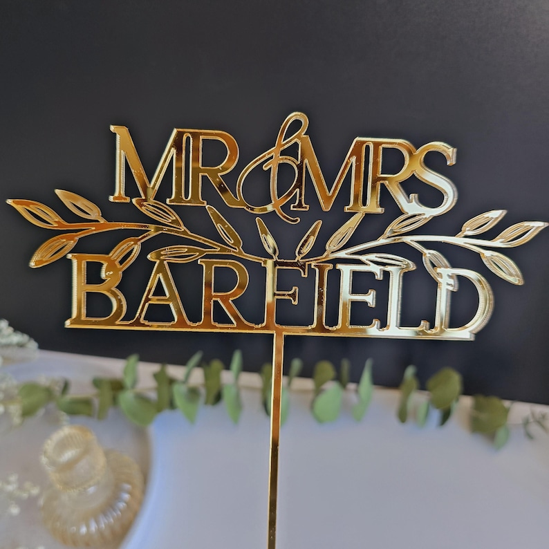 May include: Gold mirror acrylic cake topper with the text "MR & MRS BAREFIELD" and a leafy design.