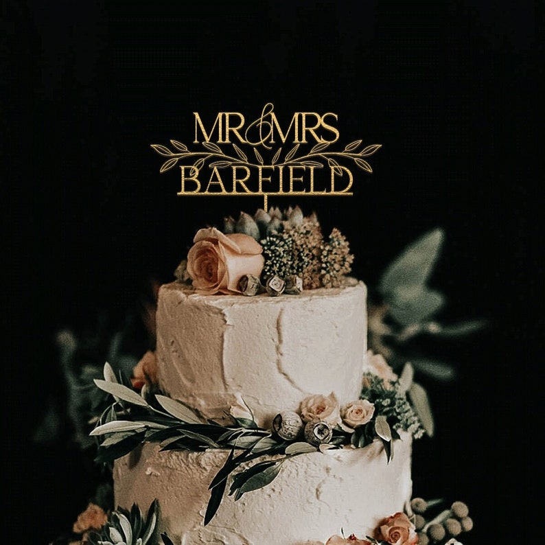 May include: A two-tiered wedding cake decorated with greenery and flowers. The cake topper is a gold sign that reads "MR & MRS BARFIELD".