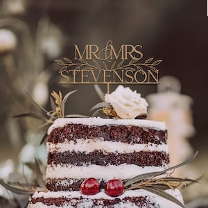 May include: A wooden cake topper with the text "MR & MRS STEVENSON" in gold lettering with a leafy design. The topper is on a two-tiered chocolate cake with white frosting and red cherries.