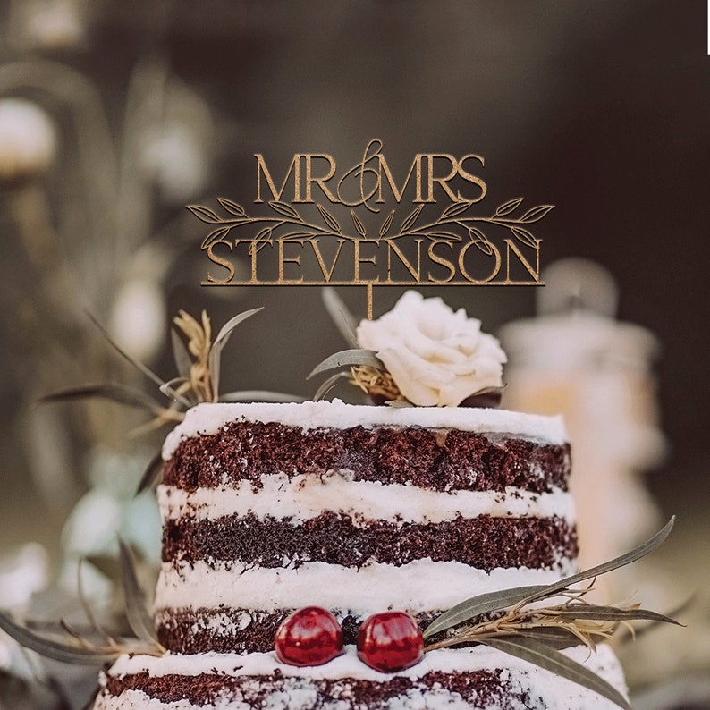 May include: A wooden cake topper with the text "MR & MRS STEVENSON" in gold lettering with a leafy design. The topper is on a two-tiered chocolate cake with white frosting and red cherries.