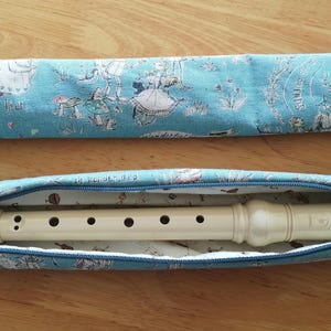May include: A blue fabric case with a zipper closure and a white recorder inside. The case has a whimsical design with a repeating pattern of dancing figures and the text "The King of the Fair".