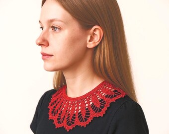 Puede incluir: Un cuello de ganchillo rojo con un intrincado patrón de hojas caladas y bordes festoneados. El accesorio se lleva alrededor del cuello sobre una camiseta negra de manga corta, mostrando un diseño de costura detallado y texturizado.