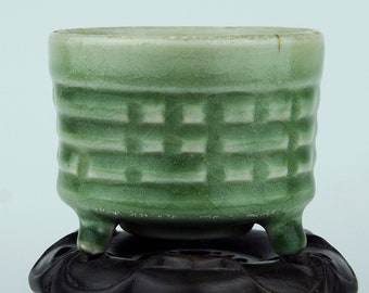 May include: A small, light green ceramic pot with a basket weave pattern, resting on a dark brown, ornate wooden stand. The pot has three small feet and a slightly textured surface.
