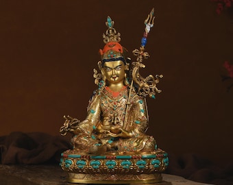 May include: A gold-toned metal statue of a seated figure adorned with turquoise and coral inlays. The figure wears an ornate crown and holds a trident staff in the left hand, resting on a lotus-shaped base decorated with blue and red stones.