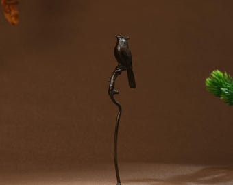 Puede incluir: Una escultura de pájaro de metal negro posada en una rama, de pie sobre una base redonda. El pájaro está mirando hacia la derecha y tiene las alas plegadas.