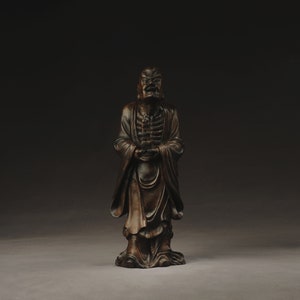 May include: A dark brown wooden sculpture of a standing figure with a stern expression. The figure is wearing a long robe and holding a small object in its hands.