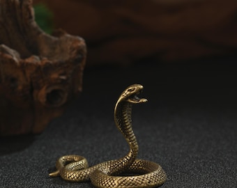 Könnte beinhalten: Eine detaillierte Cobra-Figur aus Messing, zusammengerollt mit erhobenem Kopf und offenem Maul. Die Schuppen der Schlange sind aufwendig detailliert, und das Messing hat eine warme, antike Oberfläche. Die Figur steht auf einer dunklen, strukturierten Oberfläche.