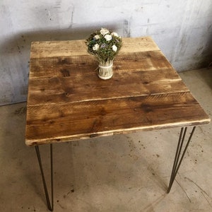 May include: A square wooden table with a rustic finish and hairpin legs. A bouquet of dried flowers sits on the table.