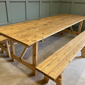 May include: A rustic wooden picnic table with two benches. The table and benches are made of natural wood with a light brown finish. The table is rectangular and has a simple design. The benches are also rectangular and have a simple design.
