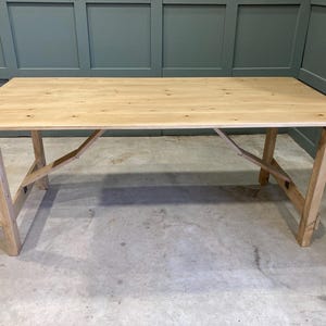 May include: A rustic wooden table with a natural finish and a simple, sturdy design. The table has a rectangular top and a trestle base.
