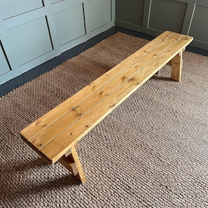 May include: A long, wooden bench with a natural finish. The bench has a simple design with a flat top and two angled legs on each end. The bench is sitting on a woven rug.