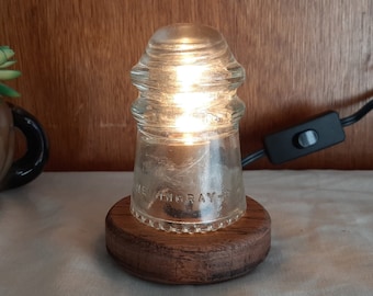 May include: A small table lamp made from a clear glass insulator with a wooden base. The lamp is lit and has a black cord with a switch.