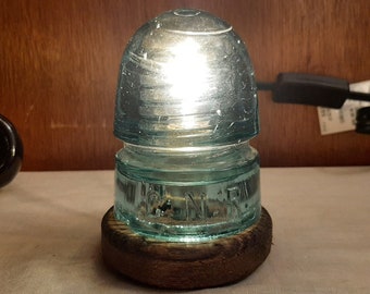 May include: A vintage glass insulator lamp with a light bulb inside. The insulator is a light blue color and has the text "24 N.R." on the side. The lamp is sitting on a wooden base.
