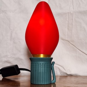 May include: A large, red Christmas light bulb with a teal base and gold trim. The bulb is lit and has a classic teardrop shape. A black power cord extends from the base. The light is on a wooden surface.