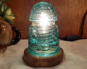 May include: A teal blue glass insulator lamp with a wooden base. The lamp is lit and casting a warm glow.
