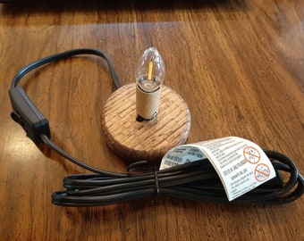 May include: A small wooden lamp base with a clear light bulb and a black cord with an inline switch. The cord is wrapped around the base of the lamp. The lamp is on a wooden surface.