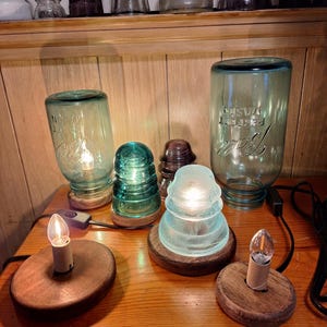 May include: Four different glass lamps with light bulbs turned on. The lamps are made from clear and aqua glass and are sitting on wooden bases. The lamps are on a wooden table.