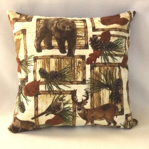 May include: A white throw pillow with a brown, green, and beige pattern featuring a bear, deer, pine cones, and branches. The pillow has a square shape and a brown border.