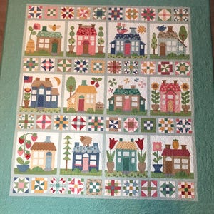 Può includere: Una trapunta con un bordo turchese e uno sfondo bianco che presenta un motivo ripetitivo di case colorate con vari dettagli, tra cui fiori, alberi e uccelli. Le case sono circondate da un bordo di quadrati di patchwork colorati.