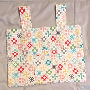 Może przedstawiać: Prostokątna torba z tkaniny z dwoma paskami. Torba ma powtarzający się wzór kolorowych geometrycznych kształtów, w tym gwiazd i diamentów, w odcieniach czerwieni, zieleni, żółci i niebieskiego na białym tle. Paski są w kolorze kremowym.