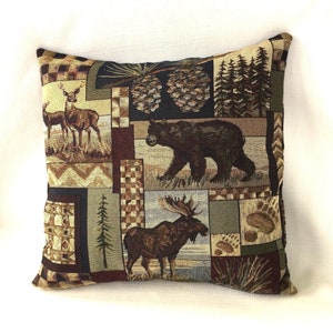 May include: A brown and tan throw pillow with a patchwork design featuring a moose, a bear, a deer, pine cones, and trees.