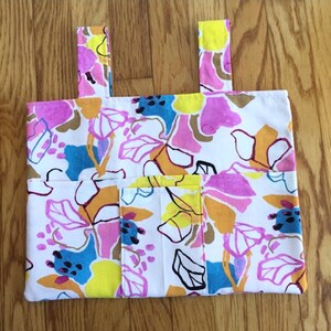 Może przedstawiać: Biała torba typu tote z żywym kwiatowym wzorem. Wzór przedstawia duże kwiaty w odcieniach różu, żółci, pomarańczy, błękitu i brązu. Torba ma dwa paski i kieszeń z przodu. Torba jest wykonana z tkaniny.