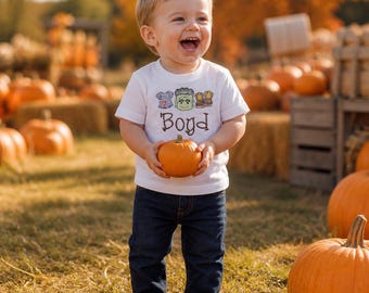 May include: A white short-sleeved t-shirt featuring a graphic of a monster head, a small shirt, and boots, with the name Boyd printed in brown text below. The shirt is paired with dark blue denim jeans, creating a casual outfit for autumn.