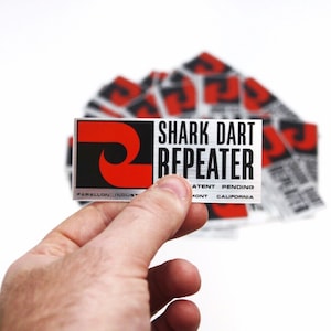 Puede incluir: Una pegatina plateada con un logotipo rojo y negro y el texto "SHARK DART REPEATER" en negro. El texto "PATENT PENDING" y "MONT CALIFORNIA" también están impresos en la pegatina.