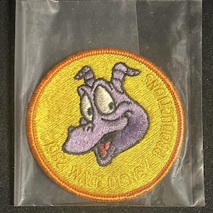 May include: A circular embroidered patch featuring a purple dragon character on a yellow background with an orange border. The text "© 1984 WALT DISNEY PRODUCTIONS" is stitched in yellow along the bottom edge of the patch, which is enclosed in a clear plastic bag.