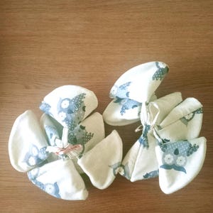 Può includere: Diversi sacchetti di stoffa bianca con un motivo a gufo blu sono disposti su una superficie di legno. I sacchetti sono legati con nastri. Il design del gufo presenta colori blu e bianchi.
