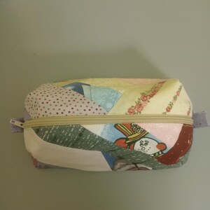 Può includere: Un astuccio rettangolare patchwork con cerniera e vari motivi di tessuto, tra cui floreali, pois e forme geometriche. L'astuccio ha una cerniera beige e piccole linguette di tessuto su ciascuna estremità.