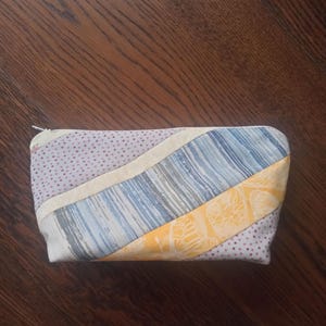 Può includere: Un astuccio patchwork fatto a mano con chiusura a cerniera. L'astuccio presenta una varietà di tessuti, tra cui pois, righe e motivi floreali nelle tonalità del blu, giallo e rosa. L'astuccio è rettangolare e sembra essere fatto di tessuto.