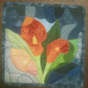 Può includere: Un arazzo da parete quadrato trapuntato con calle arancioni con foglie verdi su uno sfondo blu e grigio a motivi. I gigli hanno centri gialli. I bordi sono rifiniti con un bordo blu scuro.