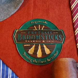 K&ouml;nnte beinhalten: Verschiedene thematische Artikel auf rotem Hintergrund: ein grauer Hut, ein Zauberstab, zwei gestreifte Krawatten, ein Holzkrug und ein Schild mit der Aufschrift "The Three Broomsticks Tavern & Inn".