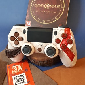 May include: A white PlayStation 4 controller with red and brown accents. The controller is sitting on a wooden base. The game case for God of War is in the background. The case is red with gold lettering and a circular design. The game is rated 18+.