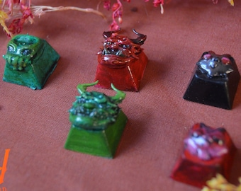 May include: Five custom mechanical keyboard keycaps featuring sculpted creature designs. The set includes a green tentacled figure, a red horned demon, a black cat-like face, a green horned monster, and a translucent red creature with pink accents.