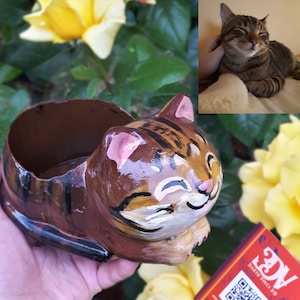 Può includere: Un vaso da fiori in ceramica marrone e nero a forma di gatto con il naso rosa e un sorriso dipinto. Il vaso è tenuto in una mano con un braccialetto colorato. Una rosa gialla è sullo sfondo. Il vaso ha un codice QR sul lato.