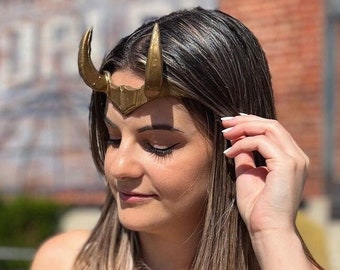 May include: A gold-colored headpiece featuring two curved horns positioned on the forehead. The accessory has a central pointed detail and a smooth metallic finish. It is worn across the brow, resting above the eyes, and is crafted with a structured, symmetrical design that curves upward on both sides.