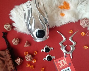 May include: A silver crown with a central black orb, a silver symmetrical ornament, and two smaller silver pieces with black centres. The set includes several beige polyhedral dice with red markings and small orange translucent crystals.